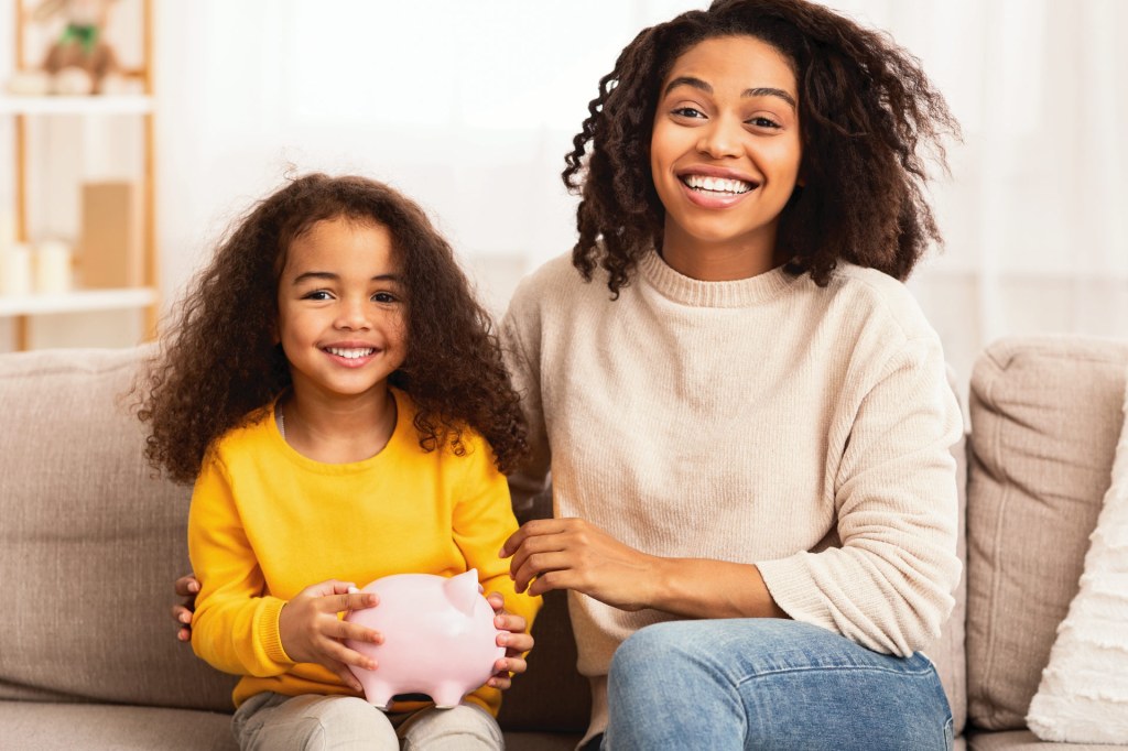 Teen and preteen African American sisters sit on a couch, holding a piggy bank