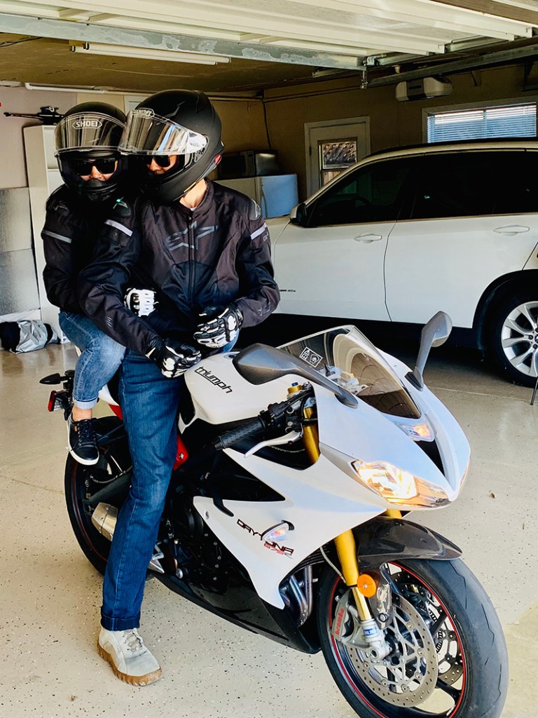 Tracy Choi and her husband David site on a white motorcycle in their garage. Each is wearing a black helmet.