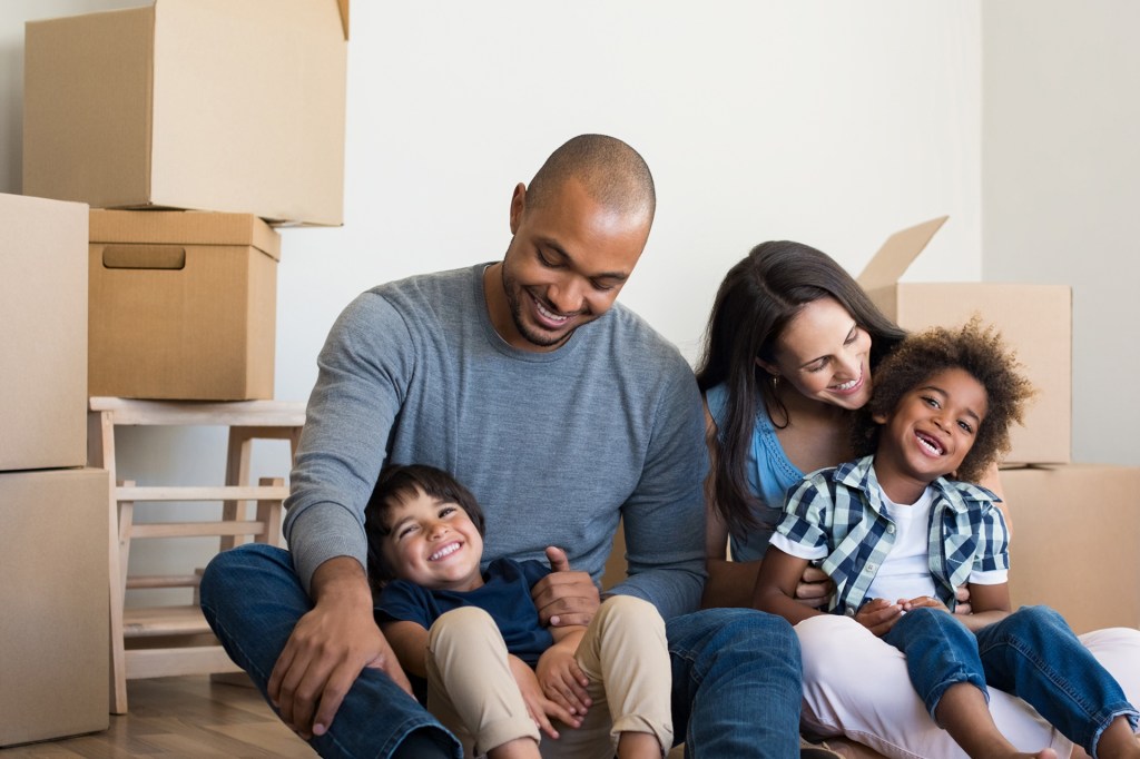 Mixed race family with mom, dad, and two kids sit on the floor of their new home with moving boxes in the background