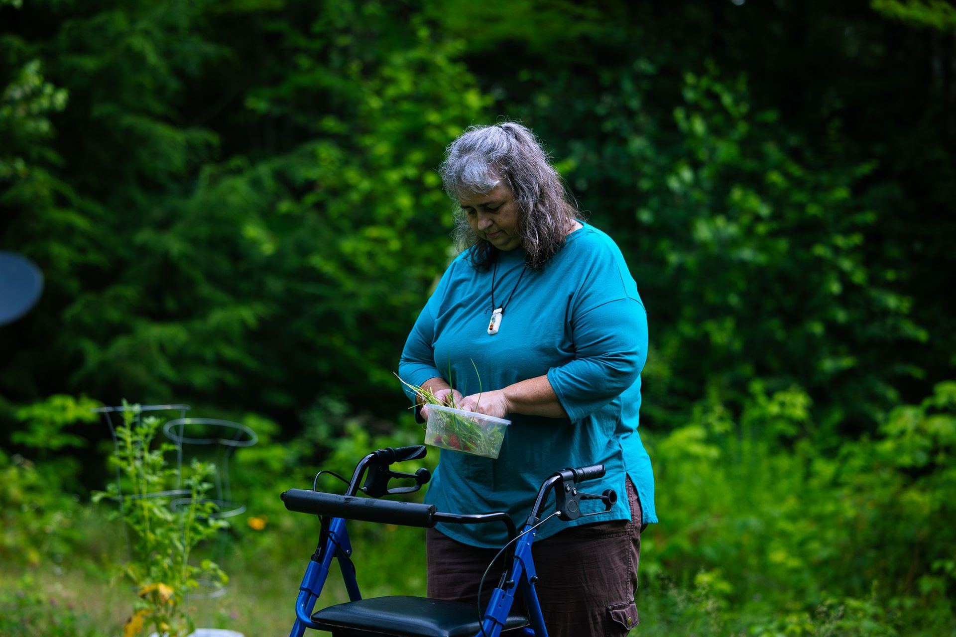 Kandie Cleaves collects vegetables in her garden. She is wearing a blue shirt and dark pants, and is using a blue walker. She wears a medical alert necklace.