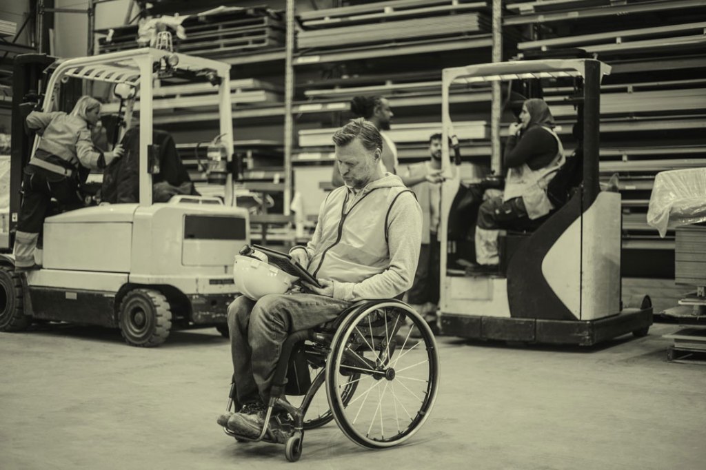 Man in wheelchair holding tablet on factory floor.