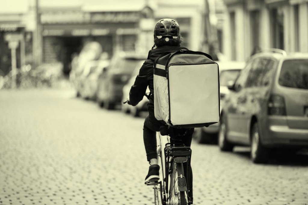 Courier person riding bicycle with thermal backpack on city street for food delivery.