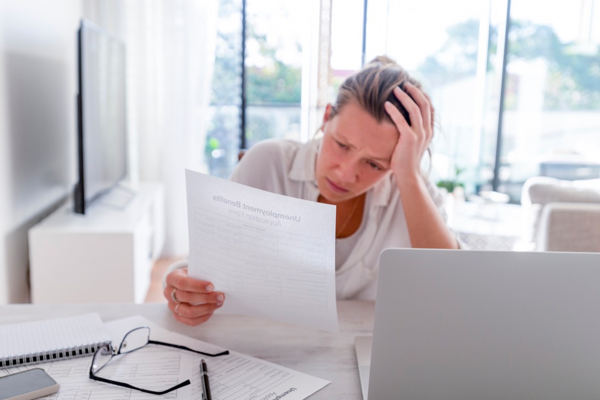Woman looking worried holding an unemployment benefits application at home. She looks very stressed and upset. She has been working from home with a living room in the background. There is a laptop computer on the table
