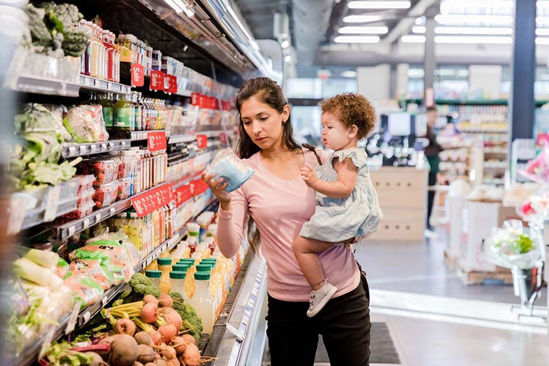 Mother holding an infant looking at prices in the produce section of the grocery store.