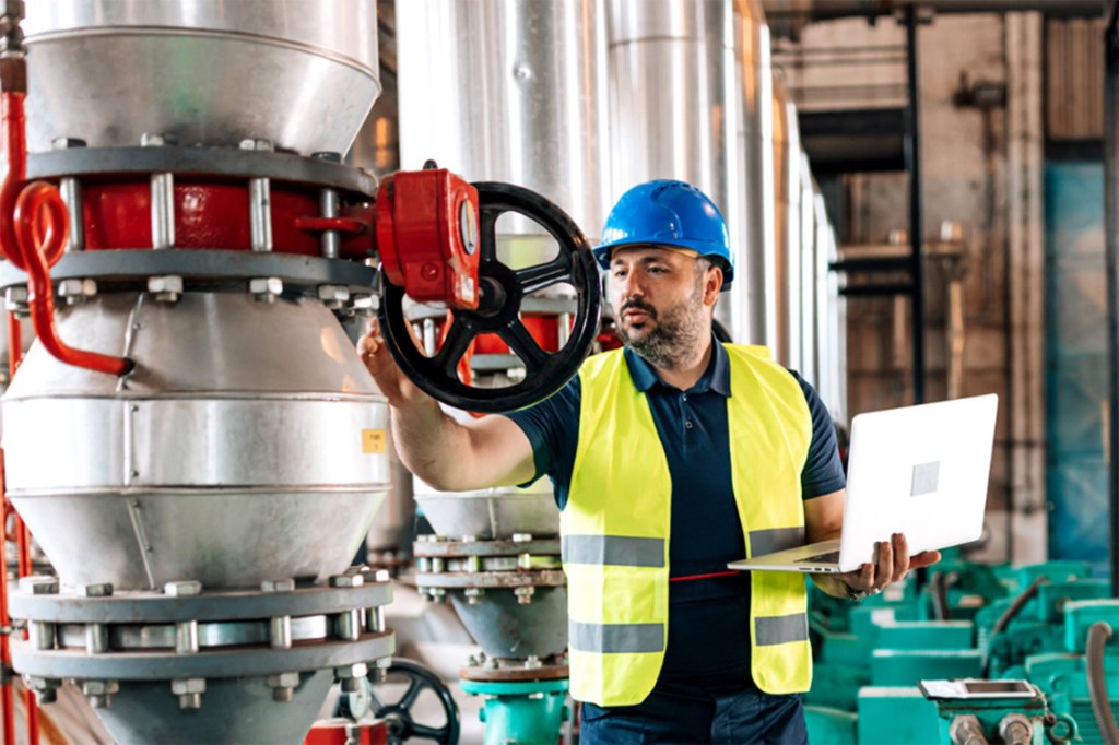 A mature Caucasian male engineer is checking a heating boiler, while holding a laptop.