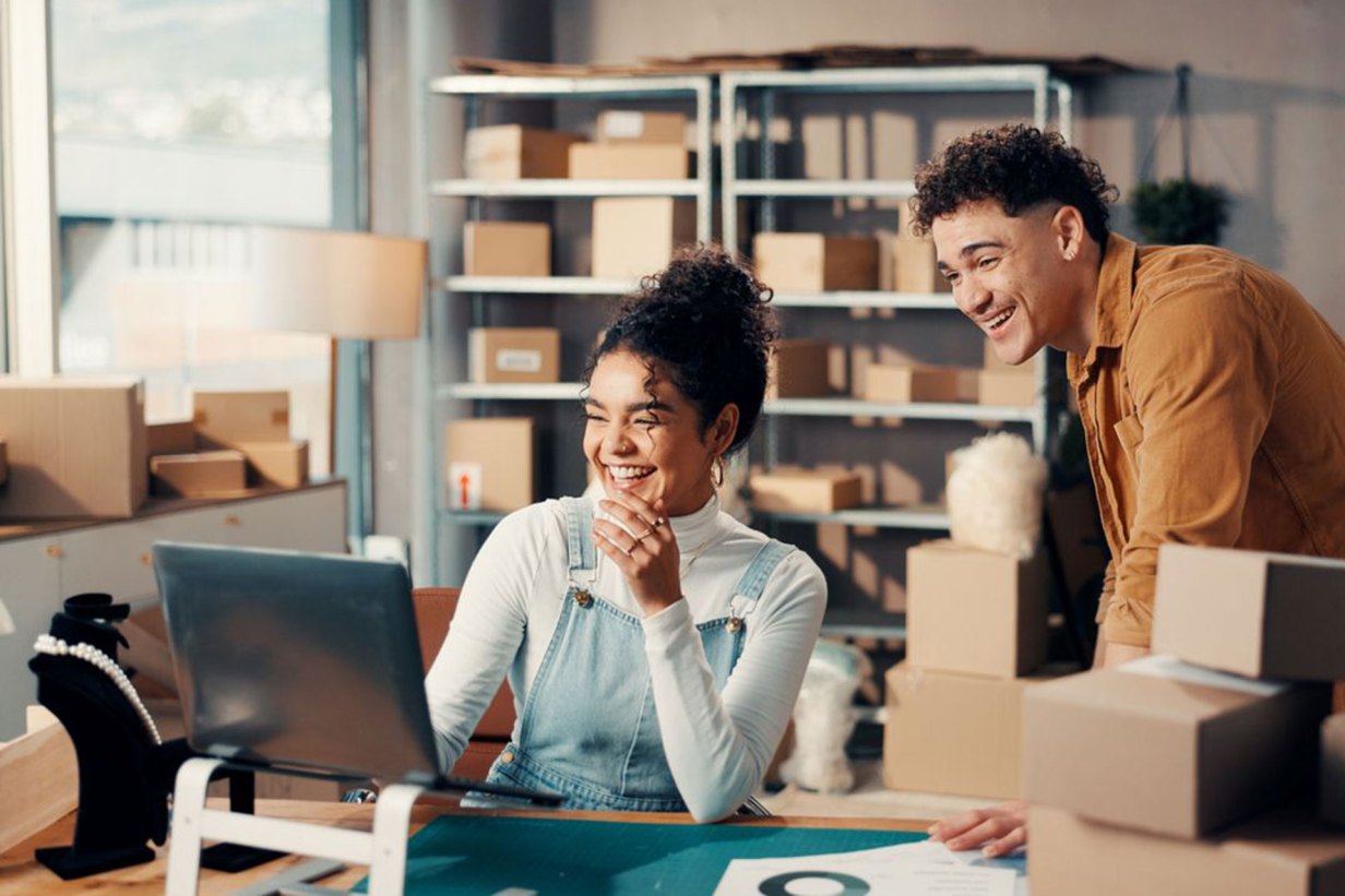 Happy, fashion and logistics team with laptop in office processing an online order with shipping boxes in the background.